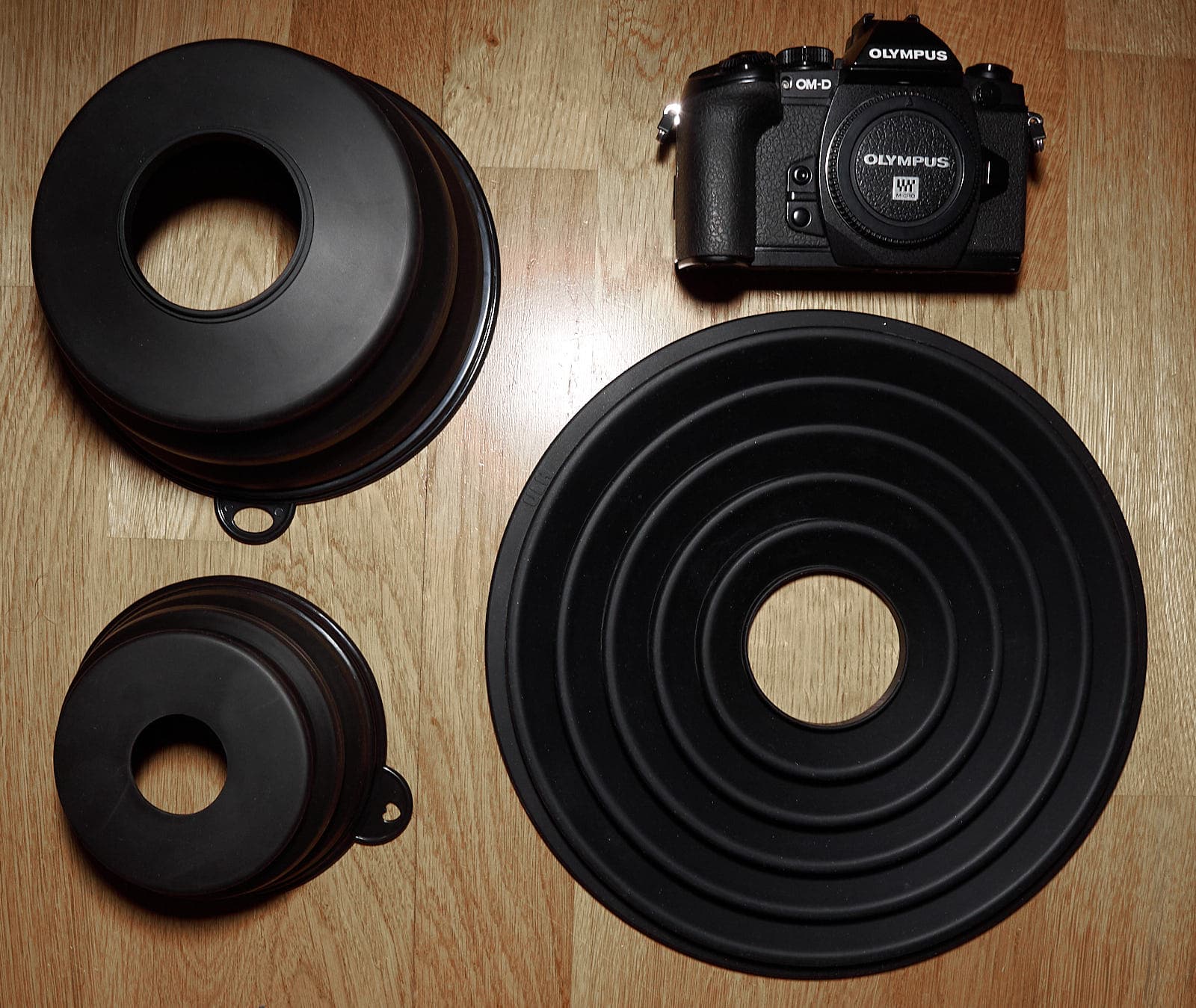 Three oversized lens hoods and a camera. On the left, the large and small lens hoods from AliExpress; on the right, an Olympus E-M1 camera and the Ultimate Lens Hood from KickStarter
